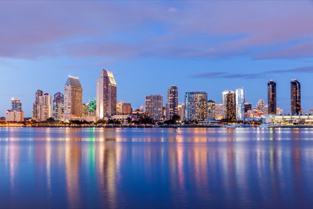 The downtown San Diego skyline glowing at dusk, its lights reflected in the calm water of San Diego Bay