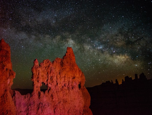 The Milky Way arching over red-lit hoodoos in Bryce's dark sky