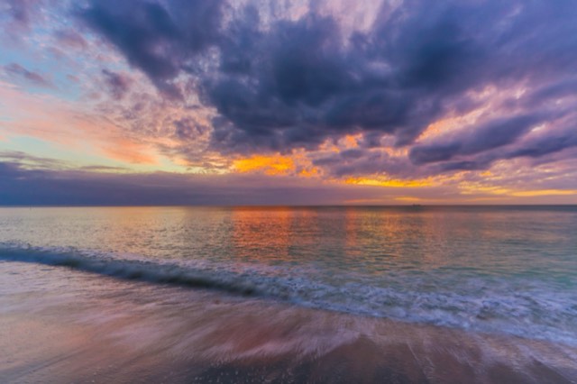 A vivid purple-and-orange sunset over the calm Gulf at Captiva Island, with a gentle wave washing the pastel-lit sand