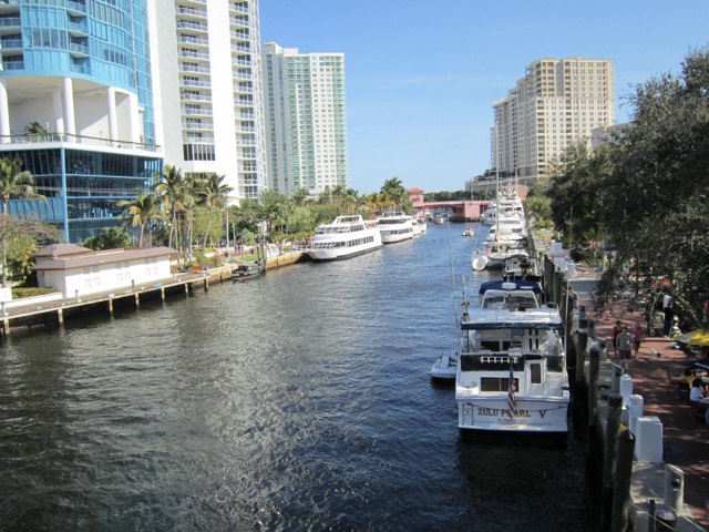 Fort Lauderdale's New River lined with palms, high-rises, and the Riverwalk, with tour boats and yachts docked along the water