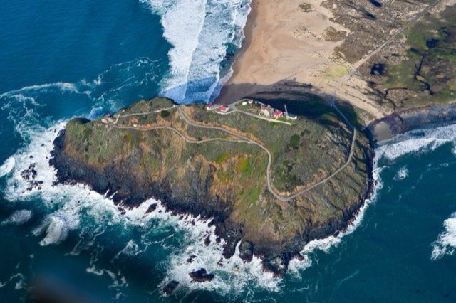 An aerial view of the Point Sur Lighthouse atop its volcanic-rock headland ringed by surf