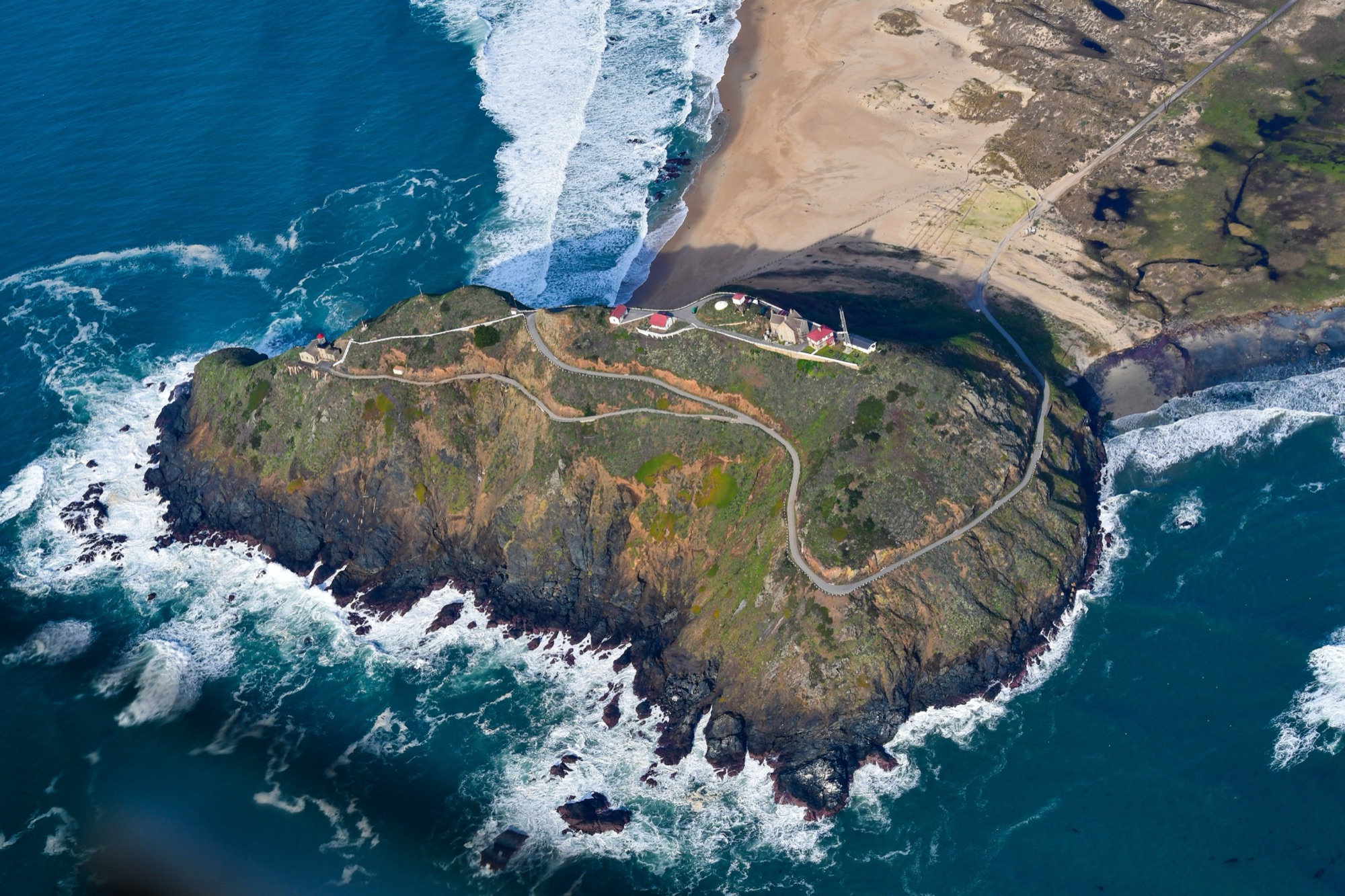 An aerial view of the Point Sur Lighthouse atop its volcanic-rock headland ringed by surf