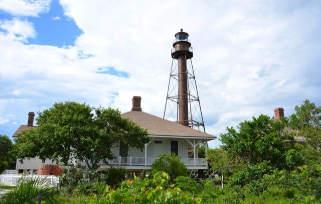 The brown iron-skeleton Sanibel Island Lighthouse standing behind its historic stilted keeper's cottage under a blue sky