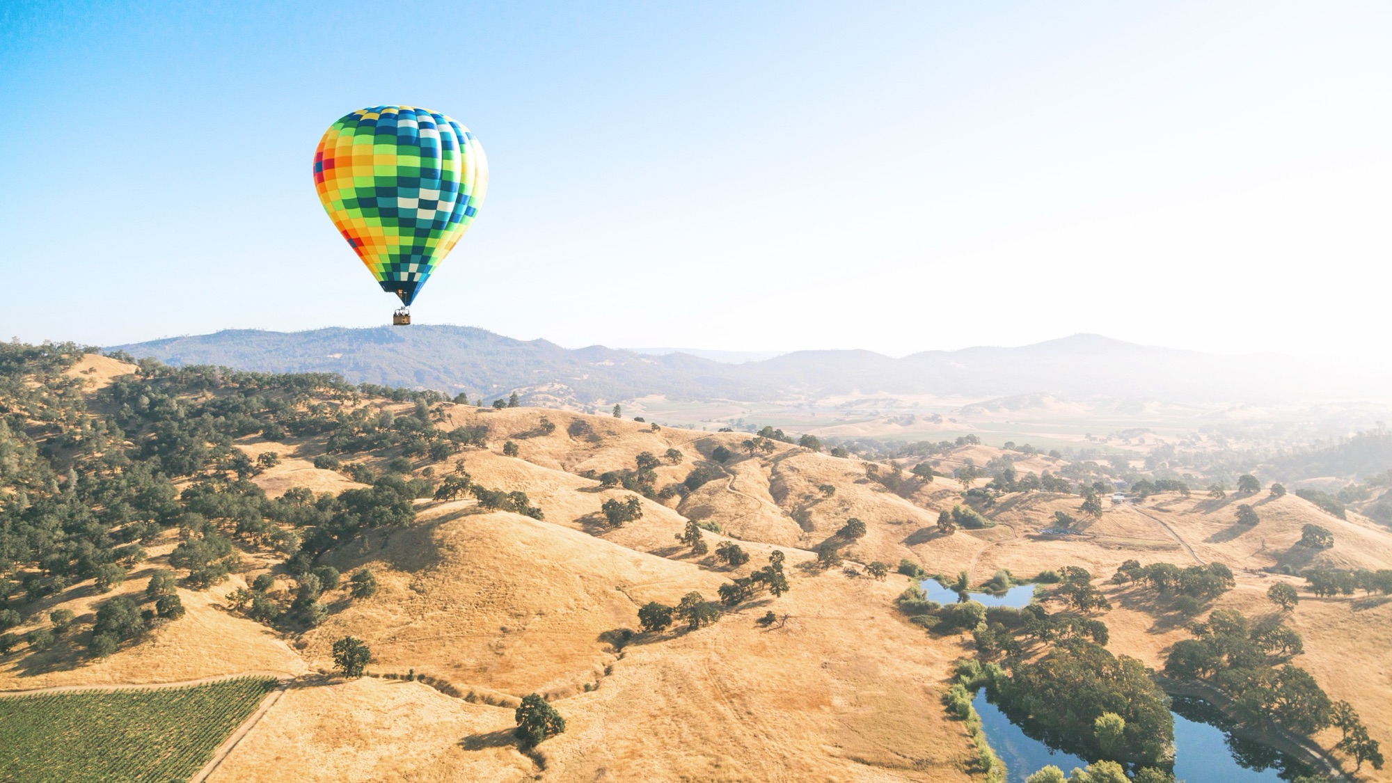 A brightly colored hot-air balloon drifting over the golden oak-studded hills and vineyards of Napa Valley at sunrise