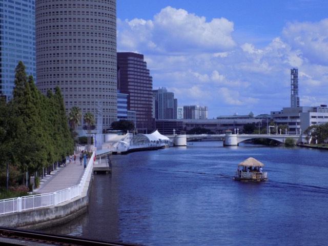 The curved Tampa Riverwalk boardwalk and its white railings following the Hillsborough River past the downtown skyline, with a tiki tour boat on the water