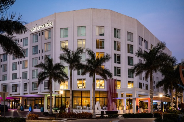 The Art Ovation Hotel at dusk — its white facade and rooftop sign lit warm above the palms on downtown Sarasota's Palm Avenue