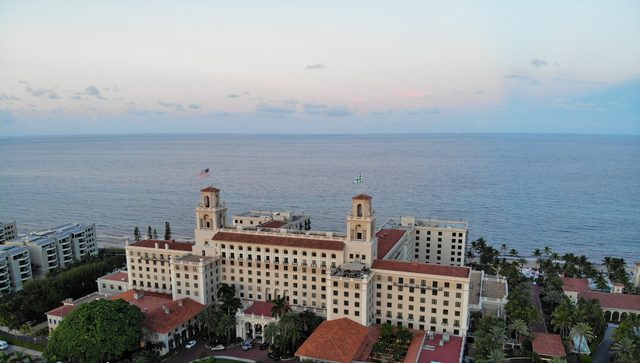 An aerial view at dusk of The Breakers resort in Palm Beach — the cream Italian-Renaissance palace with red-tile roofs and twin belvedere towers, the Atlantic Ocean stretching behind it